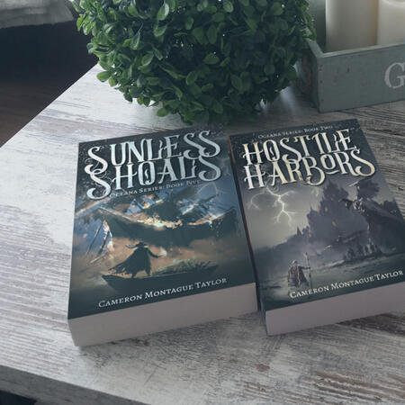Two Fantasy books lying side-by-side on a wooden table. One book is titled Sunless Shoals. The other is titled Hostile Harbors.